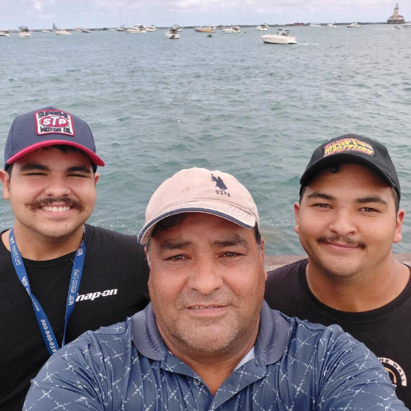 Three men smiling for a selfie by the water, with boats in the background.