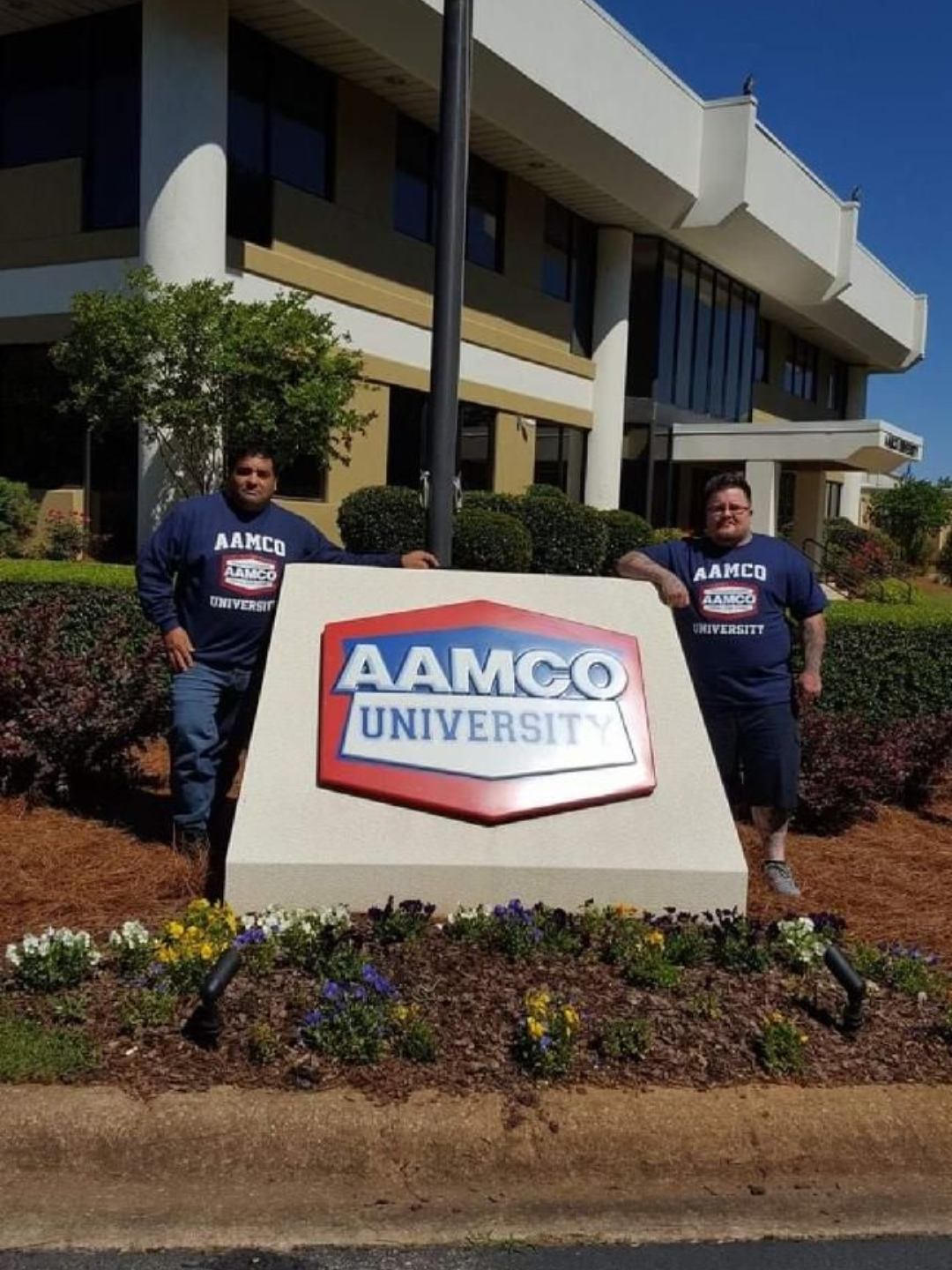 Two people standing beside an AAMCO University sign outside a building.