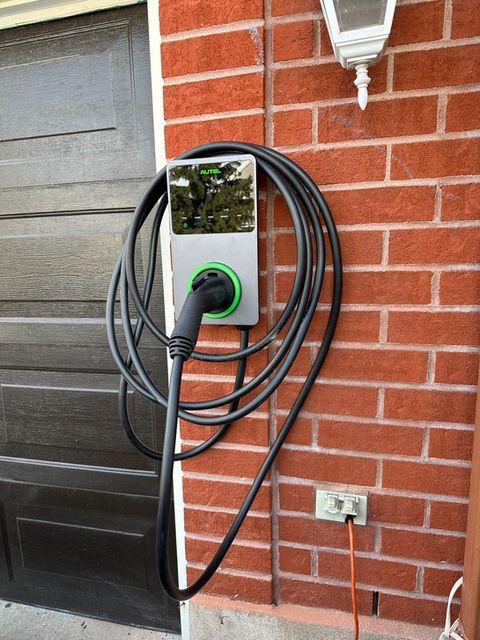 Borne de recharge pour véhicule électrique fixée sur un mur de briques, câble enroulé, branchée sur une prise.