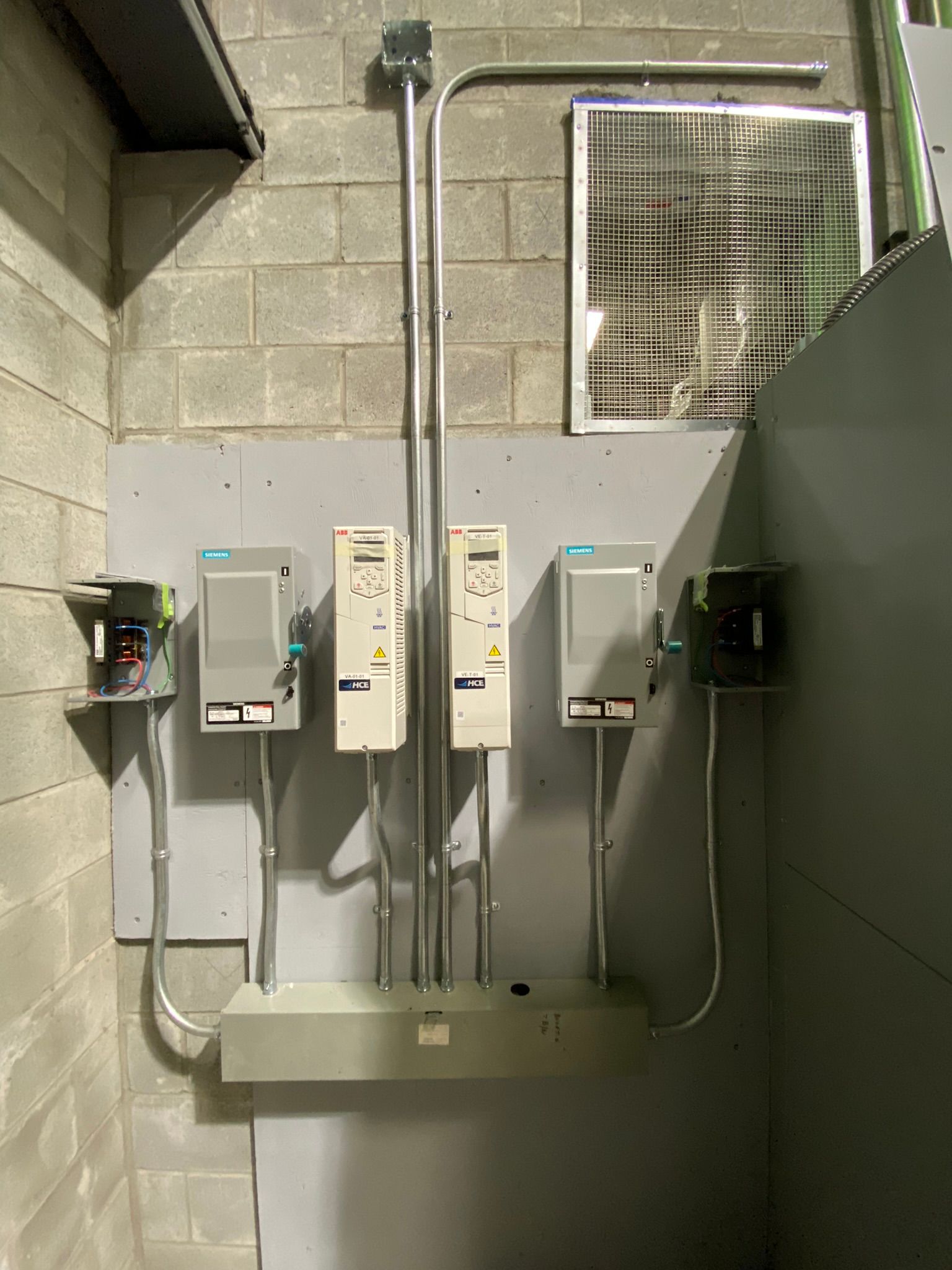 Panneaux de commande électriques fixés au mur avec conduits ; une fenêtre et un mur en parpaings gris sont visibles.
