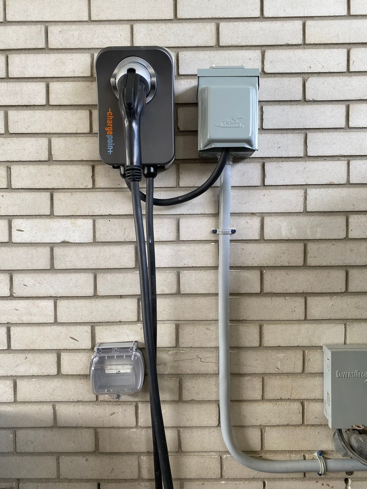 Une borne de recharge pour véhicules électriques fixée sur un mur de briques, avec un boîtier électrique et une gaine grise.