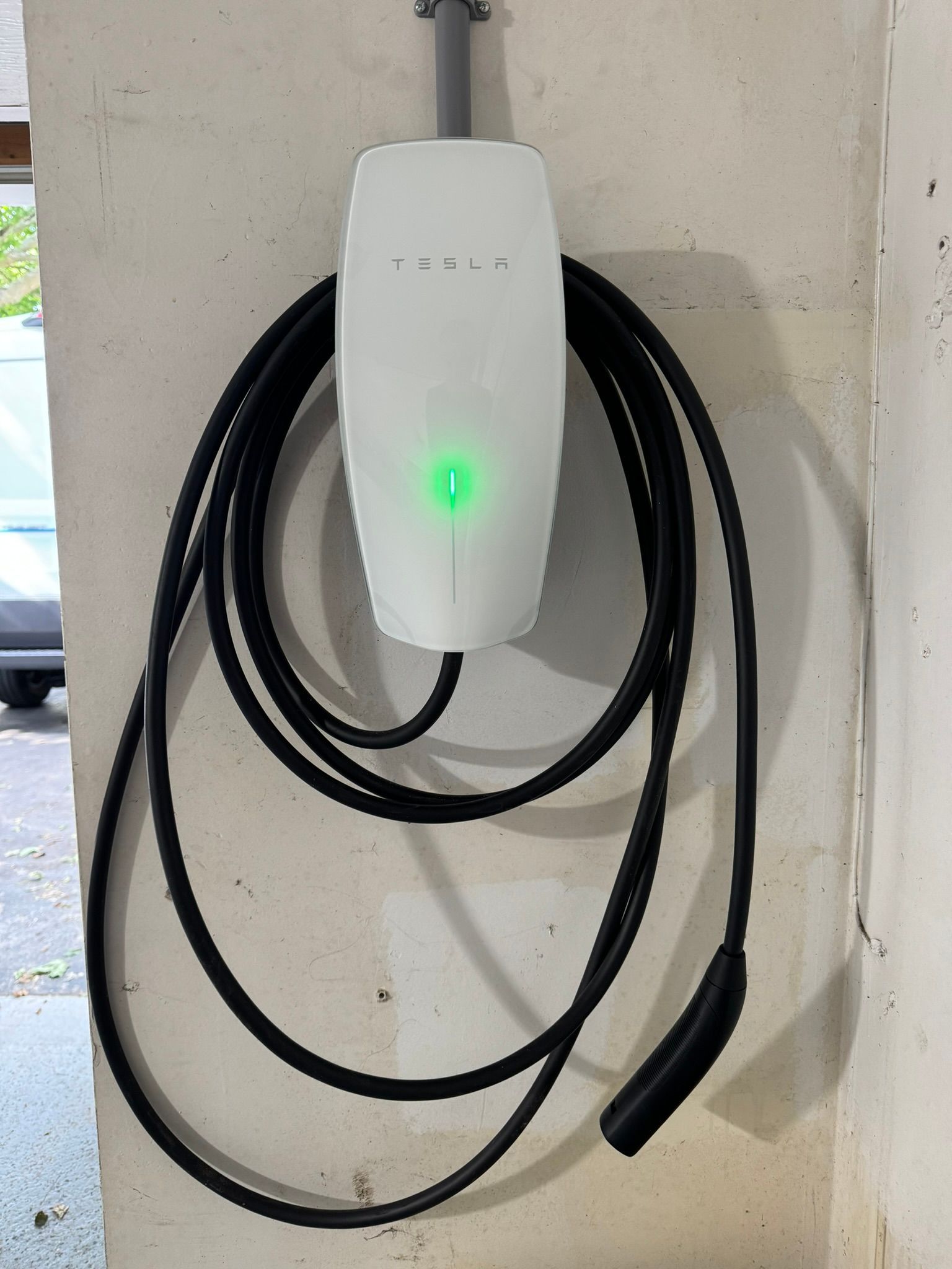 Borne de recharge blanche pour véhicules électriques, fixée sur un mur en béton, avec un câble de recharge noir enroulé en dessous.