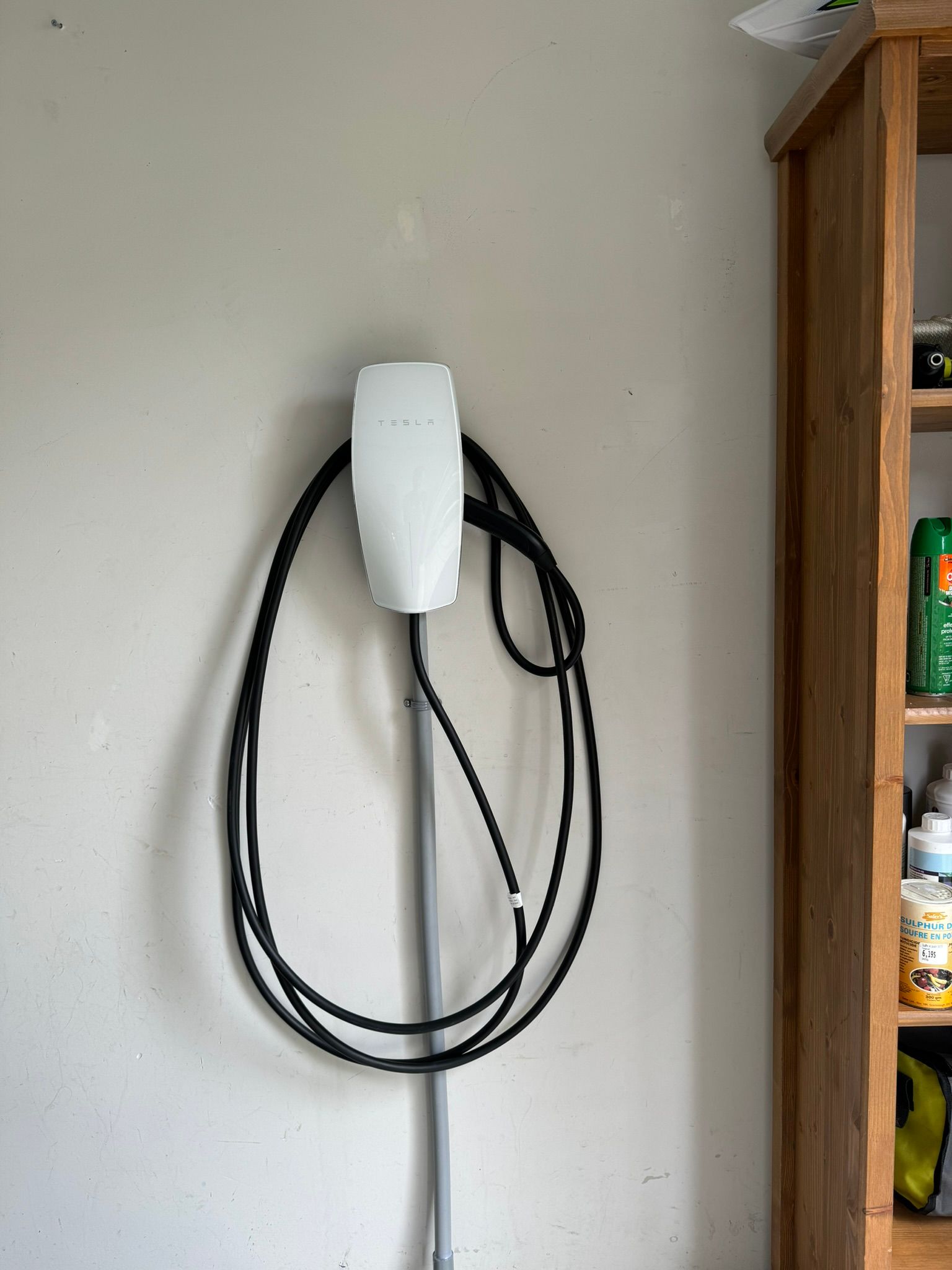 Chargeur mural Tesla blanc avec câble noir, fixé sur un mur clair, à côté d'une étagère en bois.