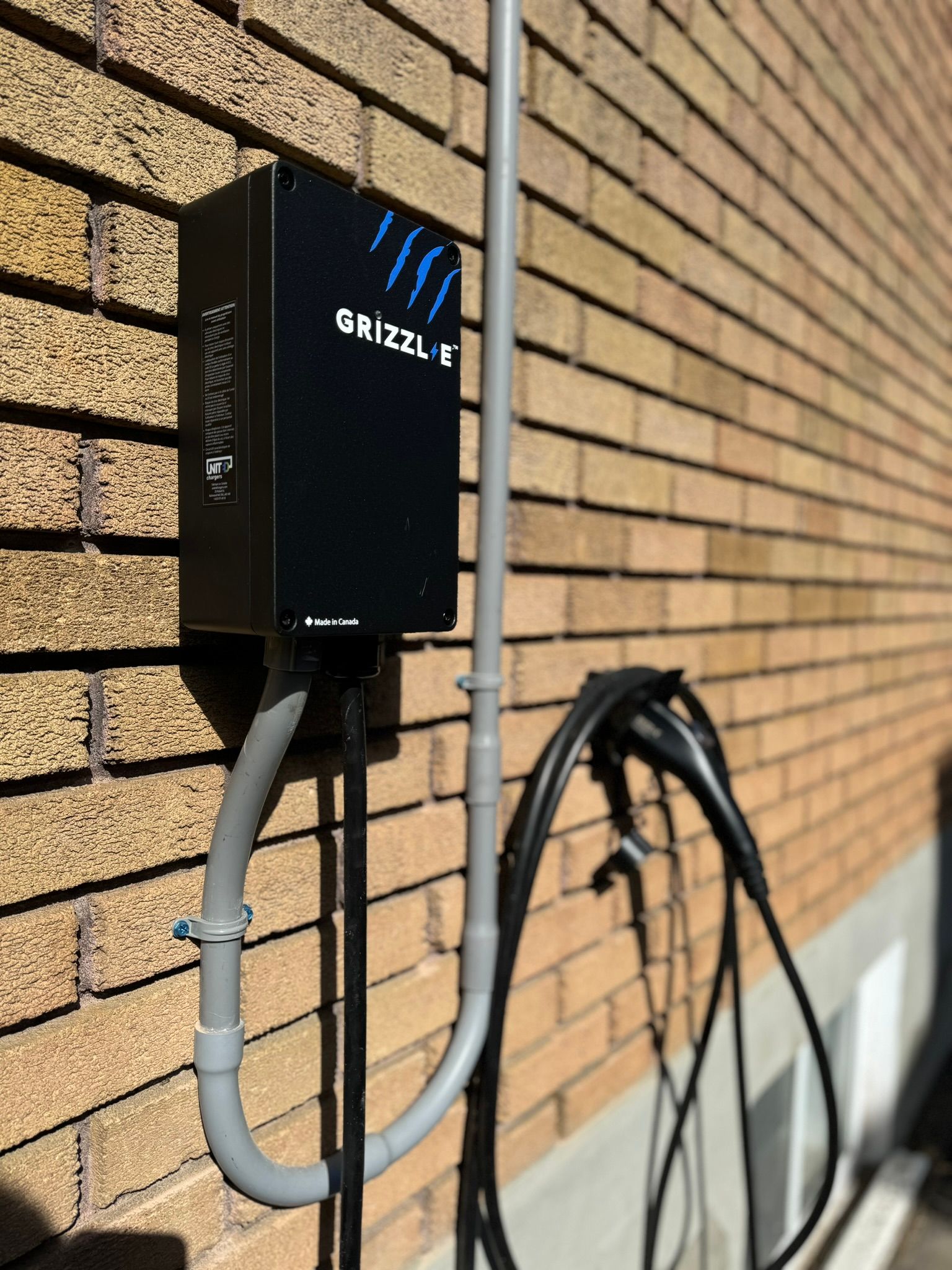 Boîtier de recharge pour véhicules électriques Black Grizzl-E monté sur un mur de briques avec conduit gris et câble de recharge suspendus en dessous.