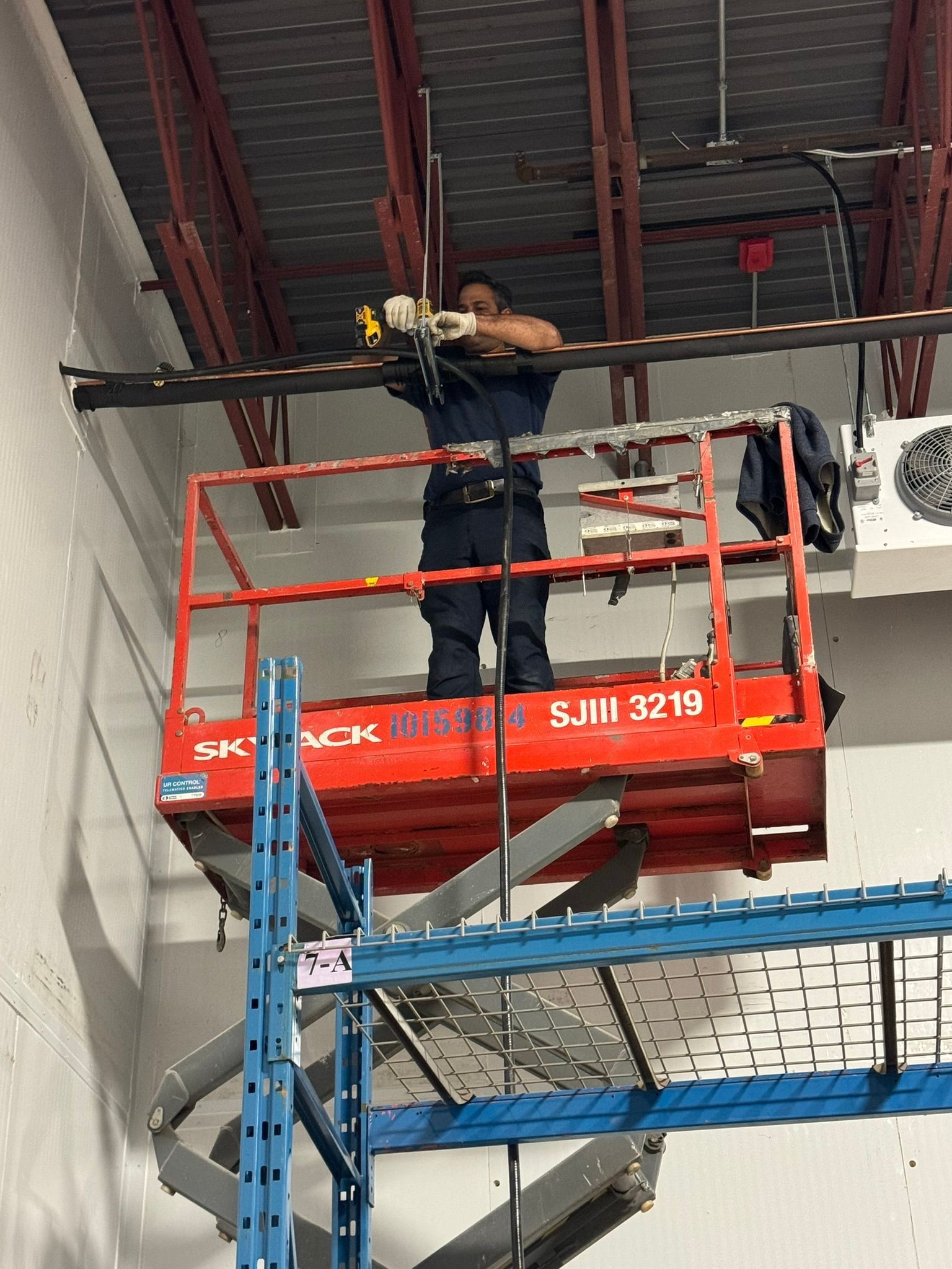 Un homme travaille sur une nacelle élévatrice à ciseaux, au sein d'une structure aérienne dans un entrepôt. Nacelle rouge, étagères métalliques au premier plan.