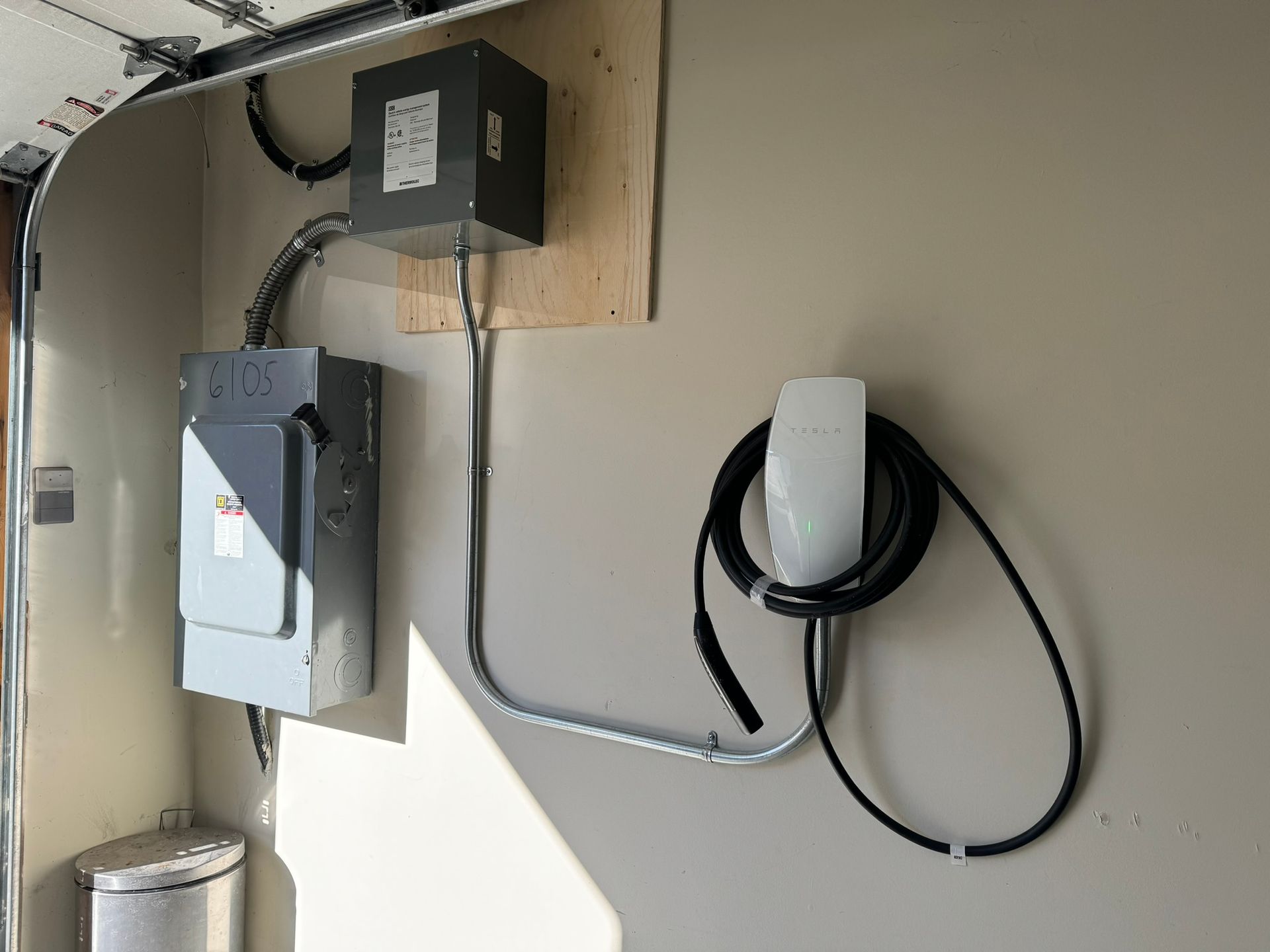 Une borne de recharge pour véhicule électrique dans un garage. Des boîtiers électriques gris, un chargeur et un câble sont fixés au mur.