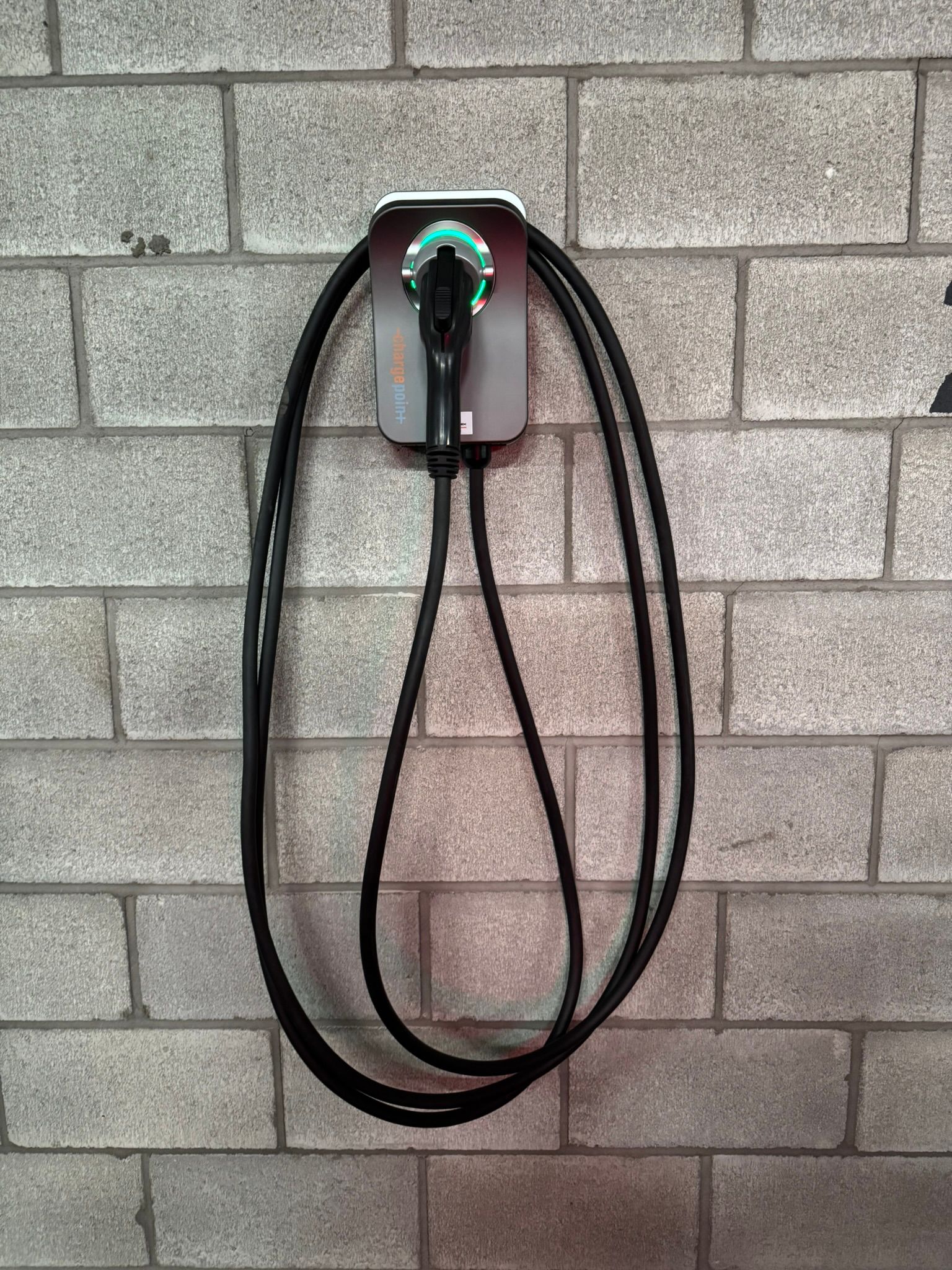 Borne de recharge pour véhicules électriques fixée sur un mur de briques grises. Un câble noir pend.