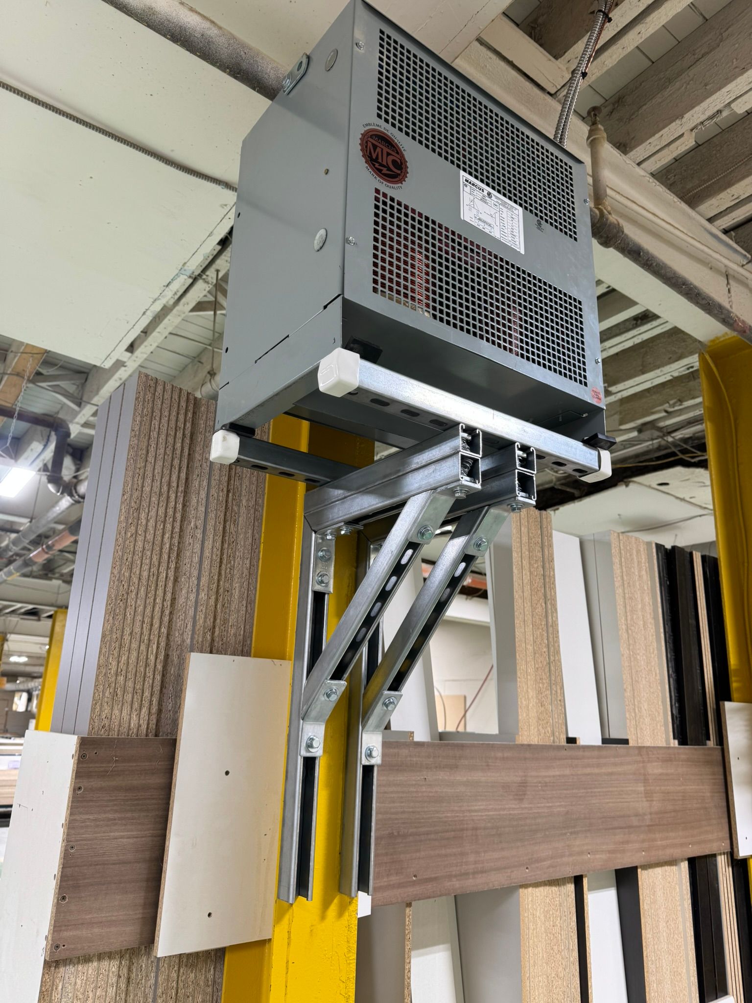 Un imposant radiateur industriel est suspendu au plafond d'une usine. L'unité grise est fixée sur un support de type ciseaux.