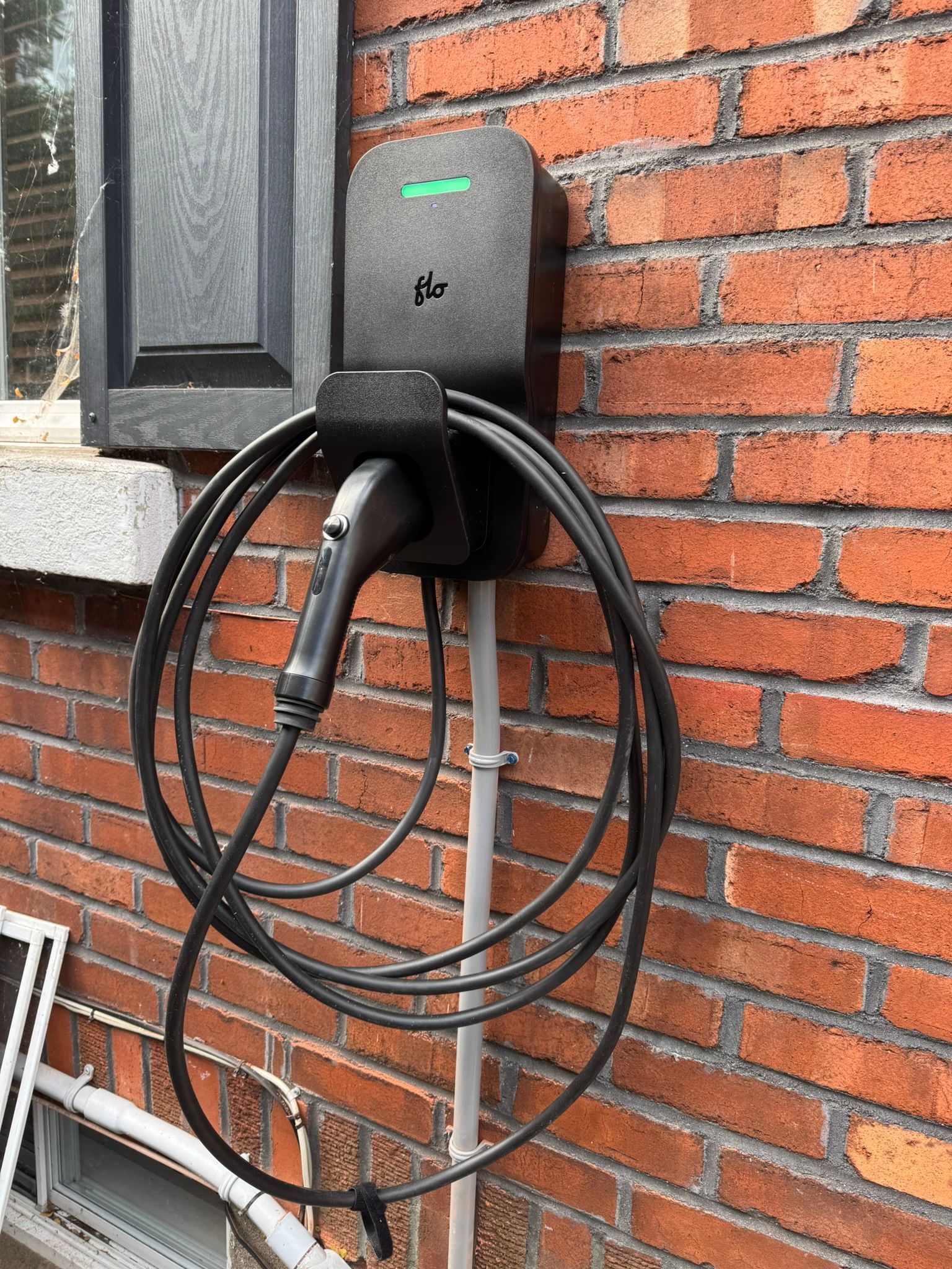 Borne de recharge noire pour véhicules électriques, fixée sur un mur de briques, avec un câble de recharge enroulé.