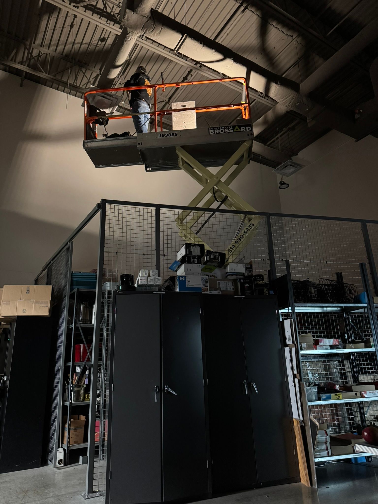 Une personne sur une nacelle élévatrice travaille au plafond d'un espace industriel avec stockage.