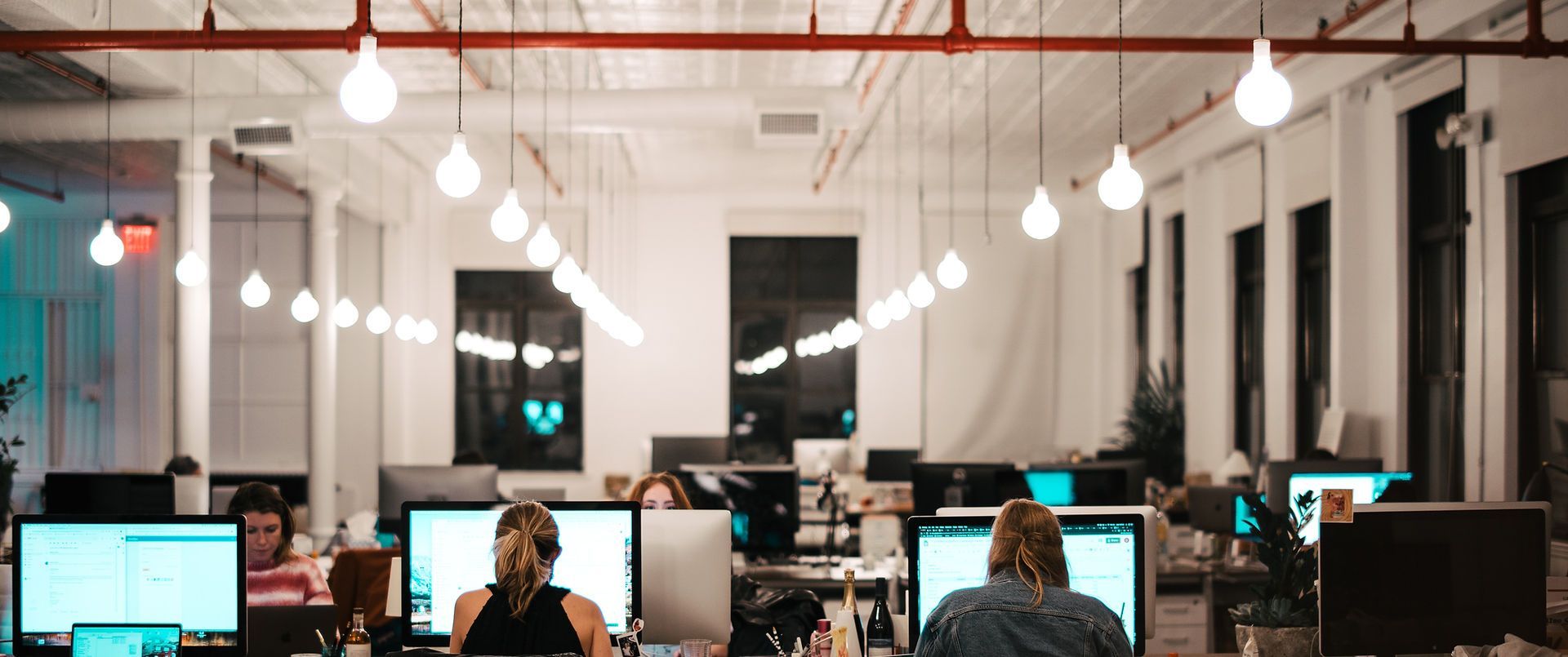 Espace de bureaux intérieur avec des personnes travaillant sur des ordinateurs sous des luminaires suspendus.