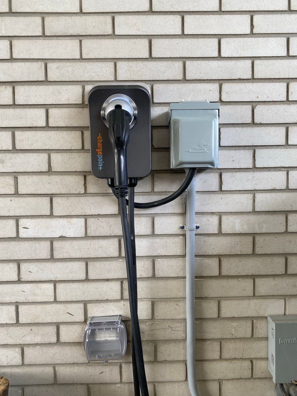 Borne de recharge pour véhicule électrique et boîtier électrique fixés sur un mur de briques. Borne et câble noirs, boîtier gris.