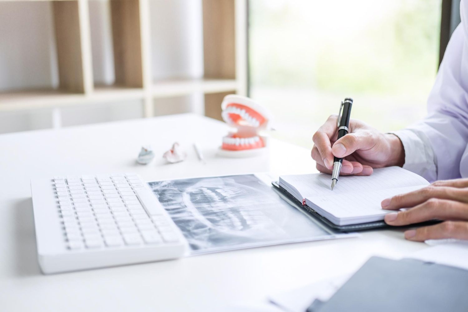 Doctor writing notes at a desk with dental X-rays, keyboard, and model teeth in a bright office