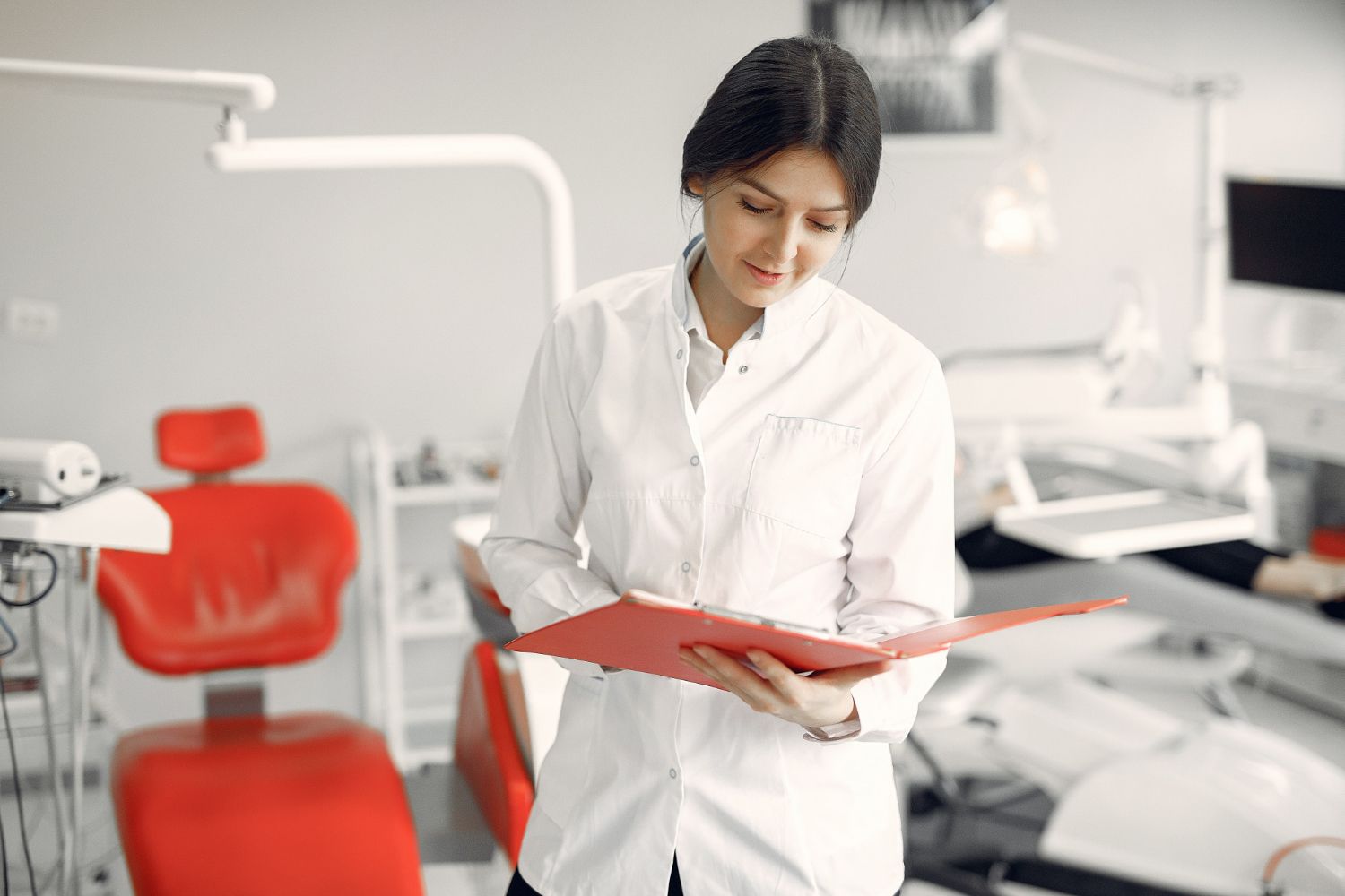 Dentist in white coat reviewing a red folder in a bright dental clinic with red chairs