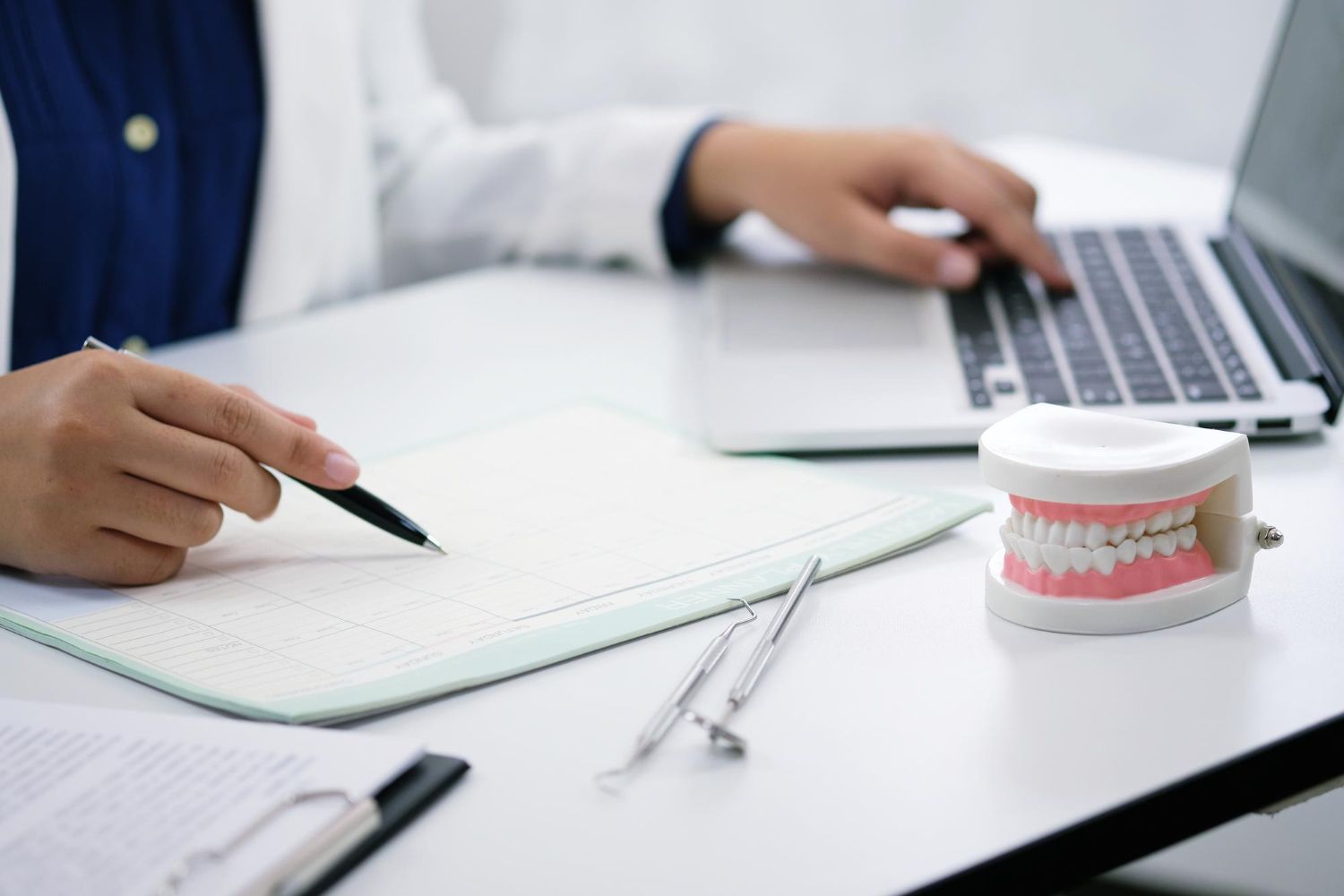 Dentist’s desk with notebook, laptop, dental tools, and a model of teeth on a white table