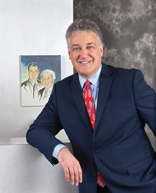 Smiling man in a navy suit with red tie, leaning against a wall beside a framed portrait.
