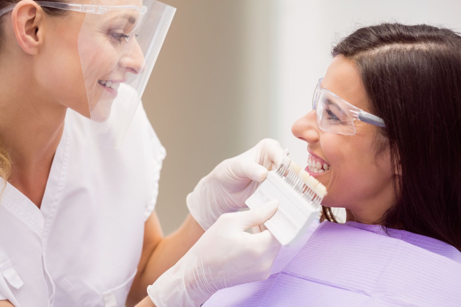 Dental hygienist cleaning a patient’s teeth while both wear protective goggles and gloves.