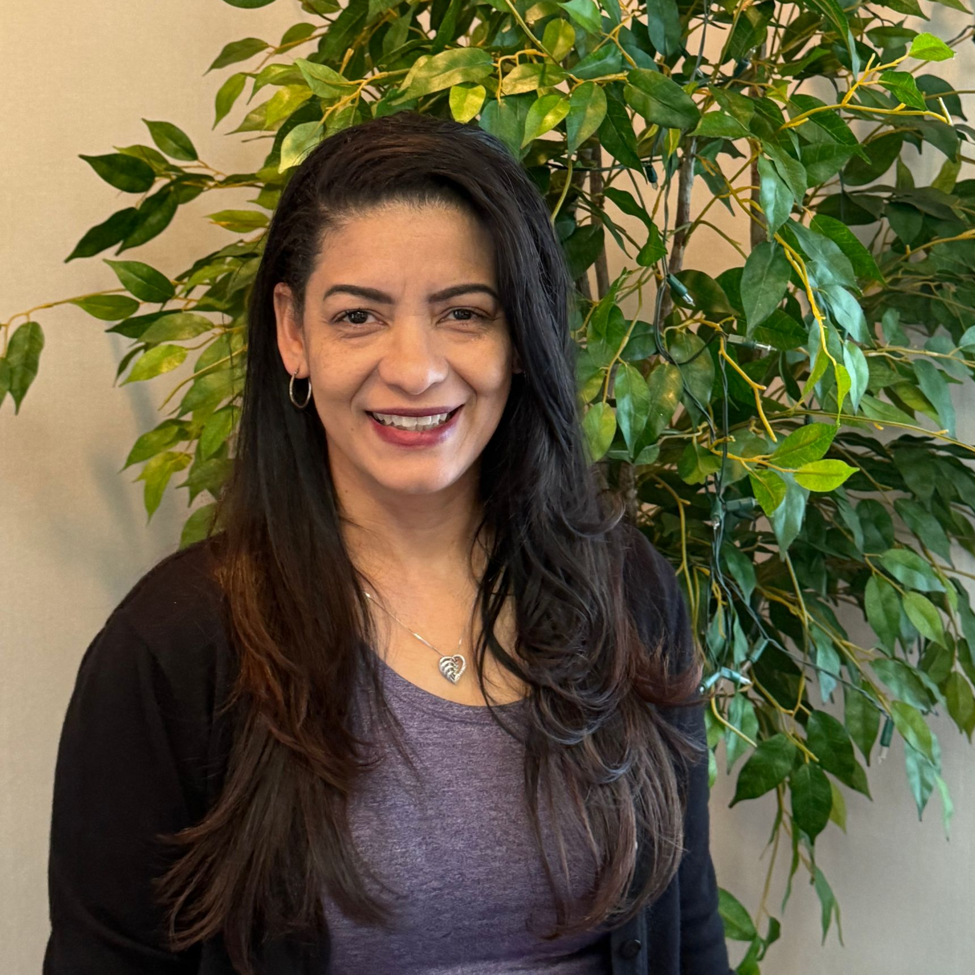 Smiling woman with long dark hair standing indoors before a leafy green plant.