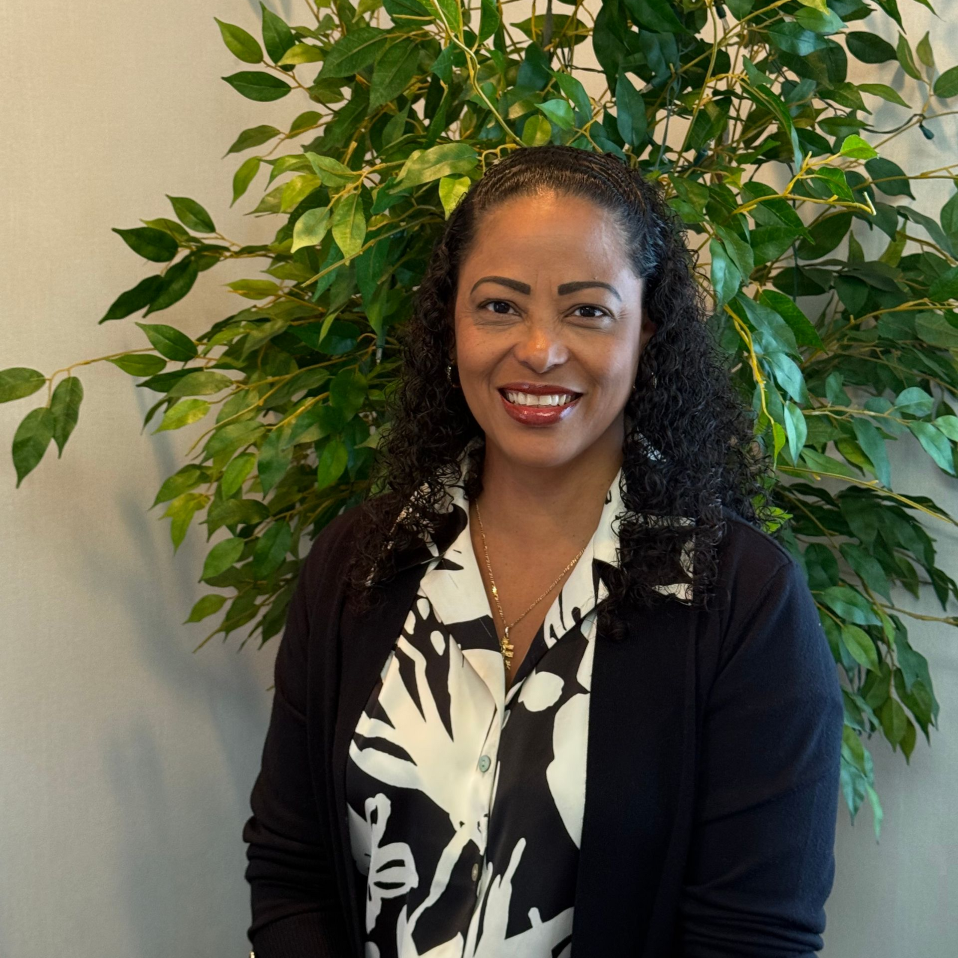 Smiling woman in a black blazer standing indoors in front of a leafy green plant.