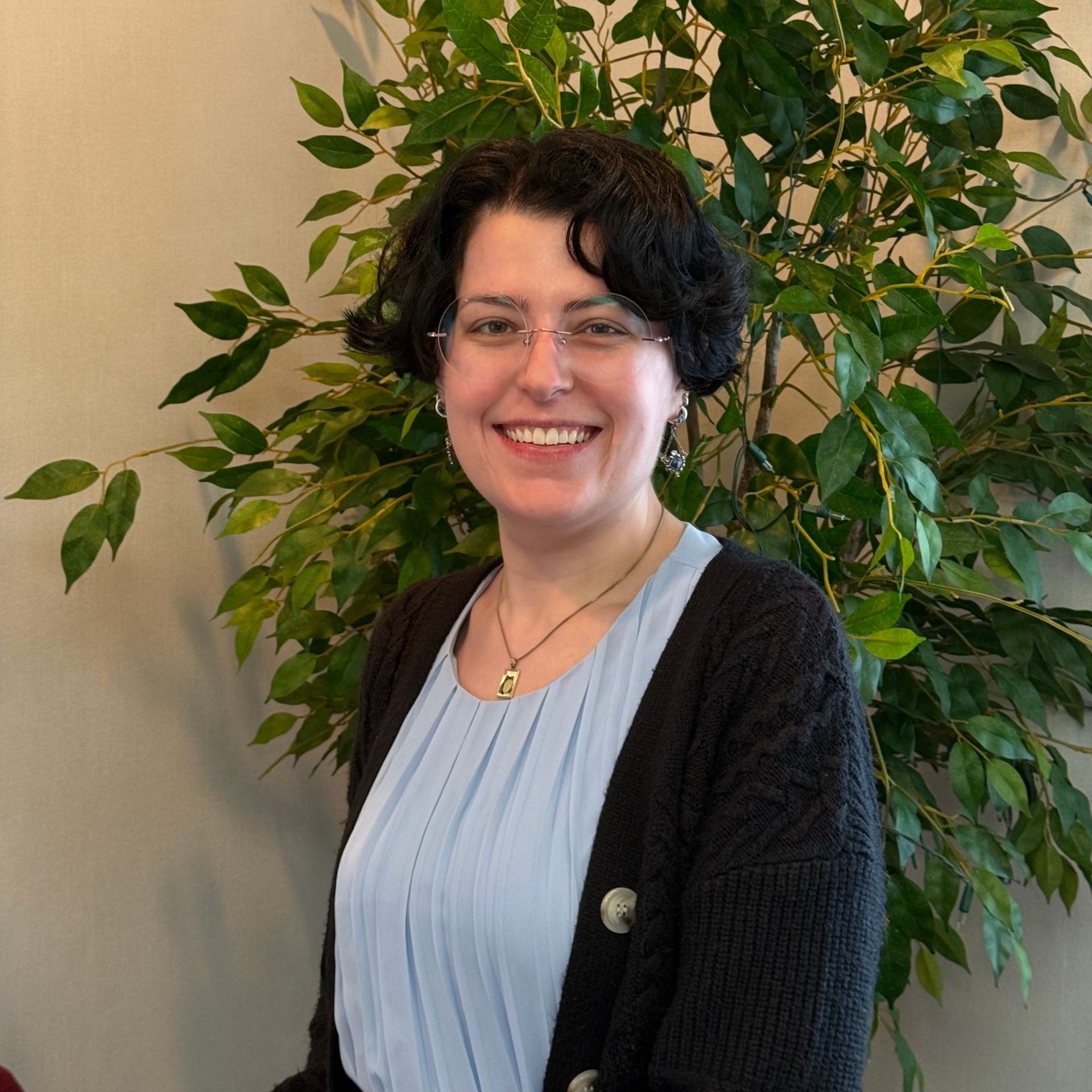 Smiling person in glasses standing indoors beside a leafy green plant.
