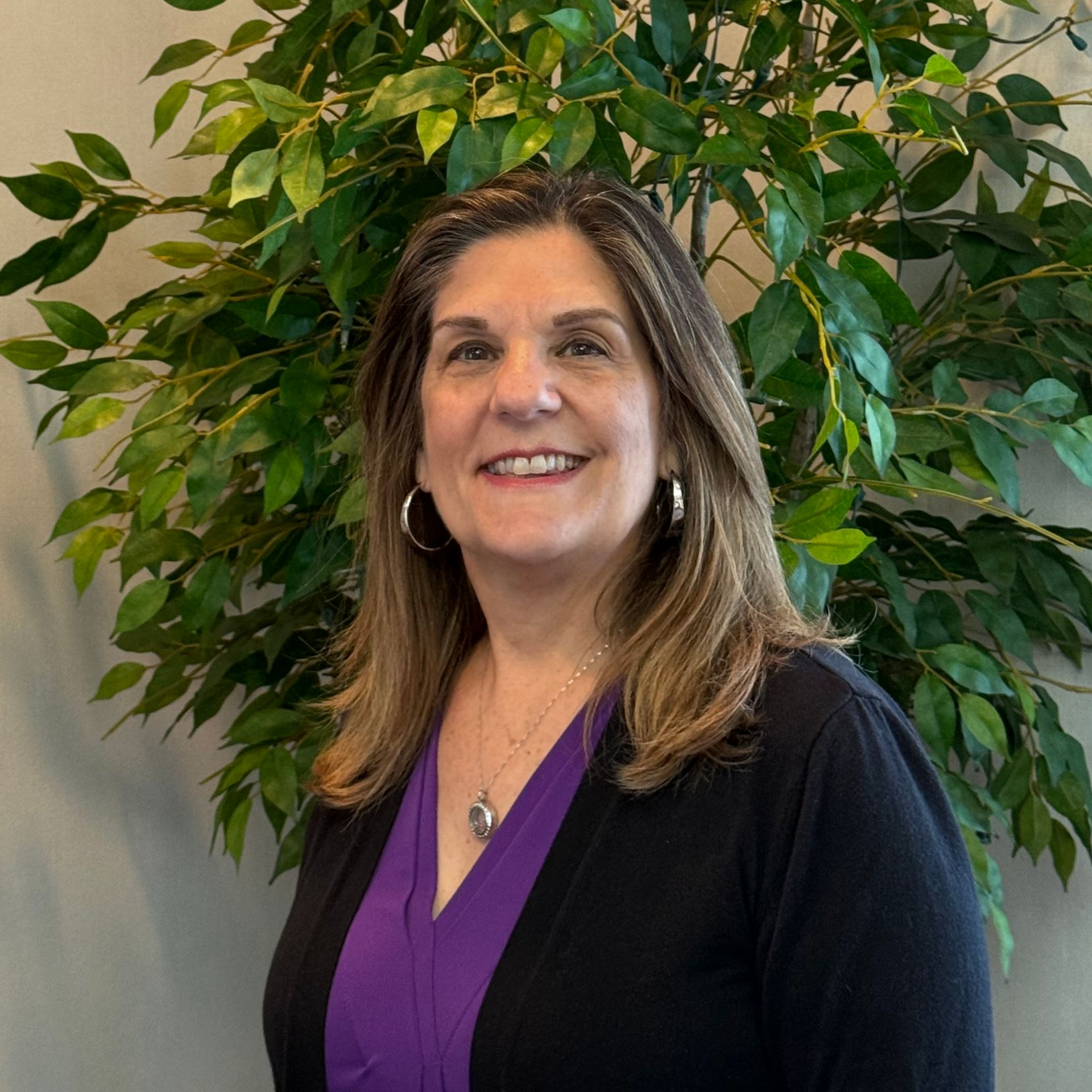 Smiling woman in a purple top and black cardigan standing before a leafy indoor plant.