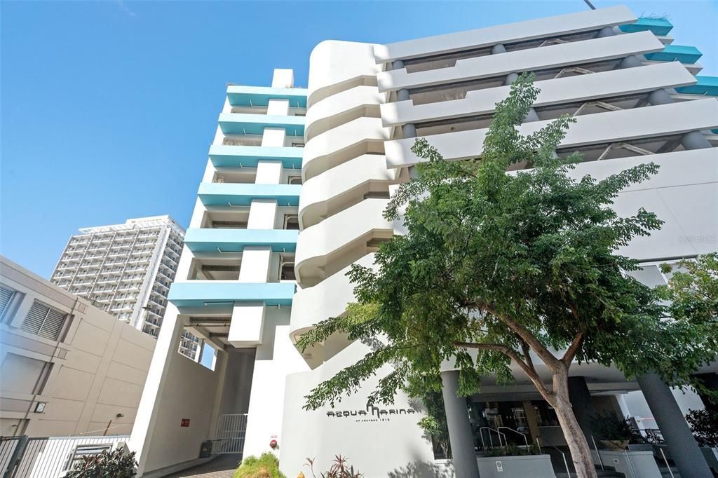 Modern white apartment building with turquoise balconies and a tree in front under a blue sky