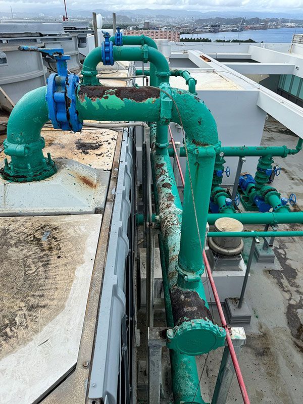 Green industrial pipes and valves on a rooftop, with city and water views in the background