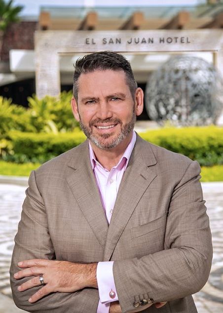 Man standing in a tan blazer in front of El San Juan Hotel, smiling with arms crossed