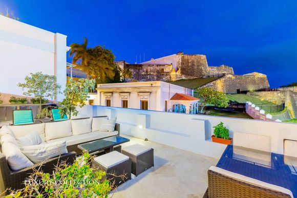 Rooftop patio with white seating, potted plants, and a lit stone fortress in the background at dusk