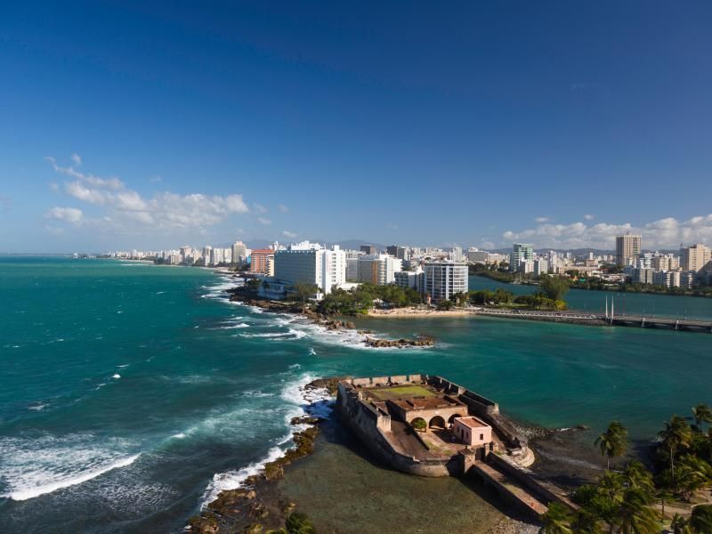 Coastal city skyline beside turquoise ocean, with a historic fort on a rocky point in the foreground