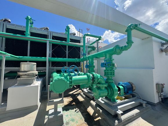Green industrial piping and machinery on a rooftop, with blue sky and clouds in the background.