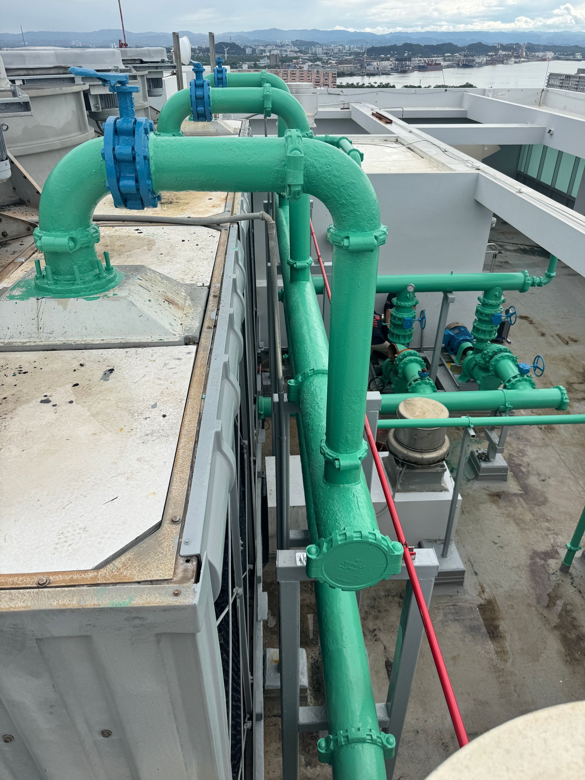 Green industrial pipes and valves on a rooftop, overlooking a city skyline.