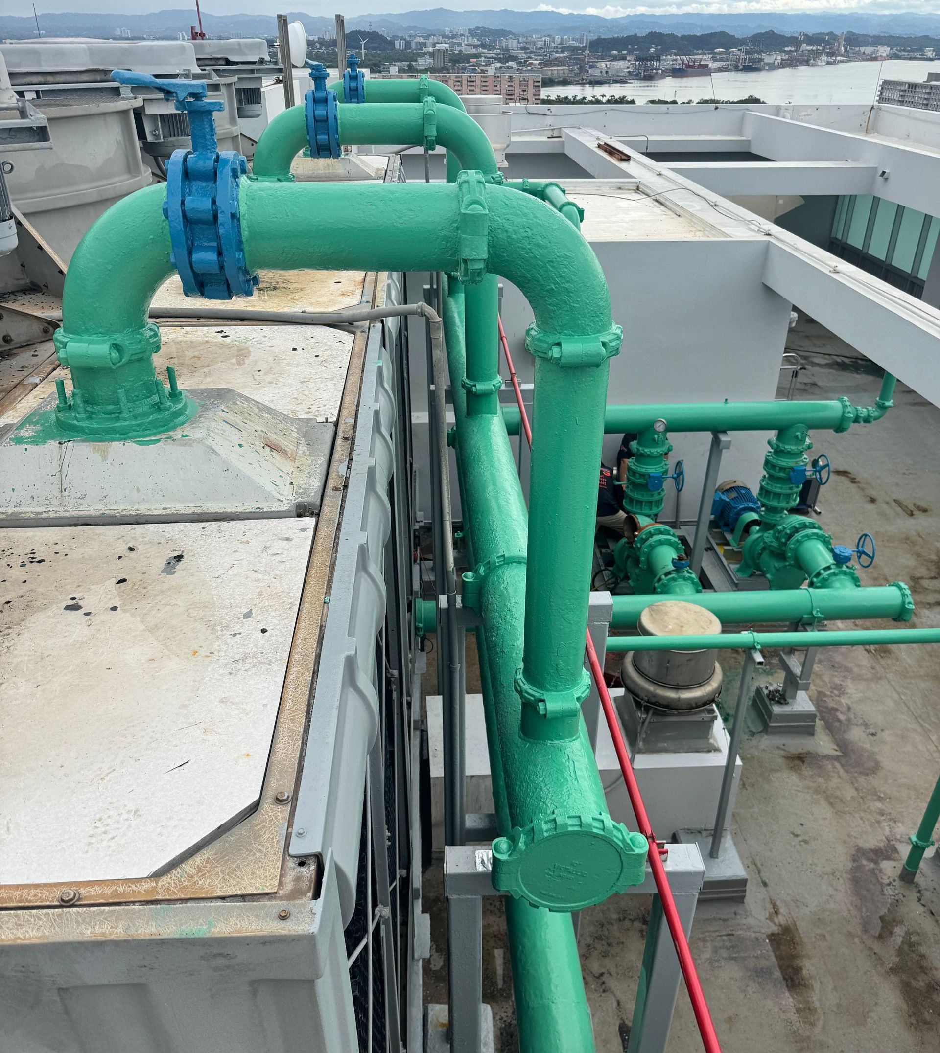 Green industrial pipes and blue valves on a rooftop, with city buildings in the background