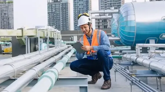 Engineer in hard hat inspecting rooftop pipes at an industrial facility, with city buildings in the background