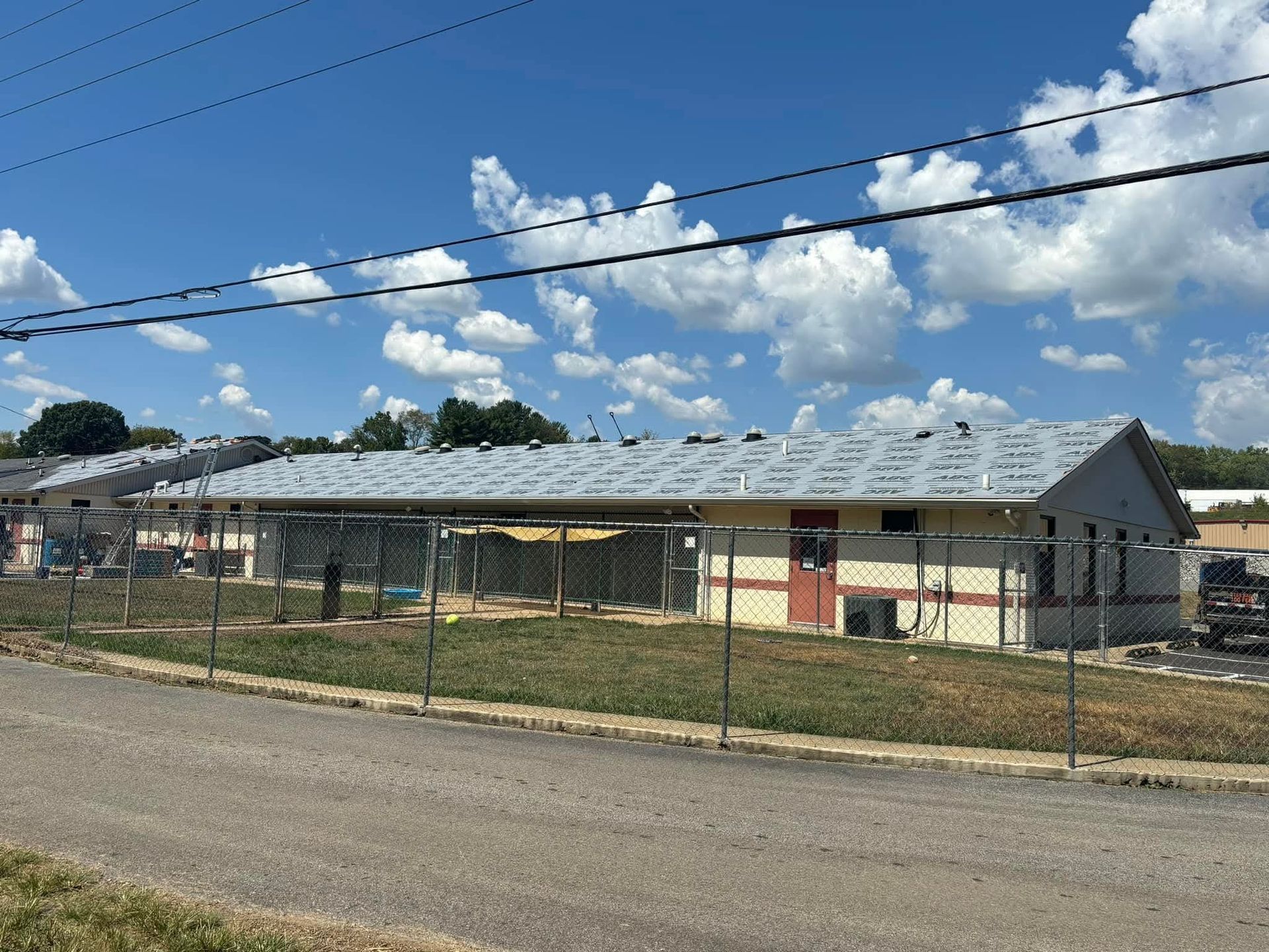 Single-story building with a fenced yard under a blue, cloudy sky beside a road