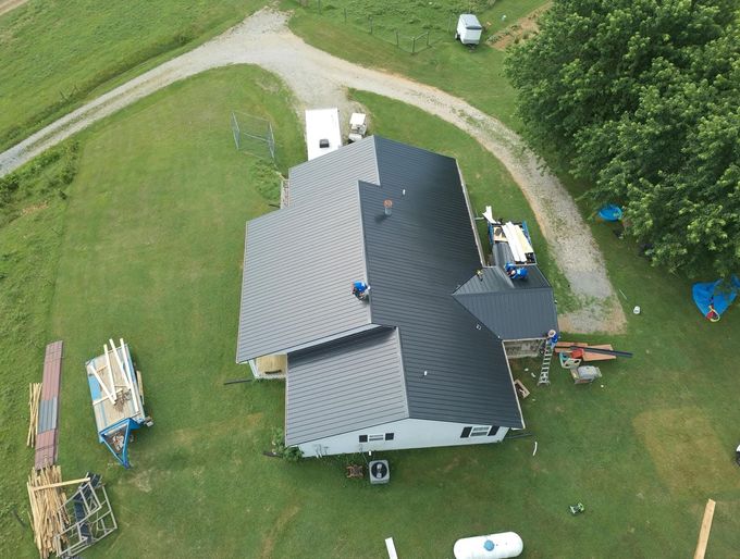 Aerial view of a gray-roofed house with lawn, trees, and scattered yard equipment.