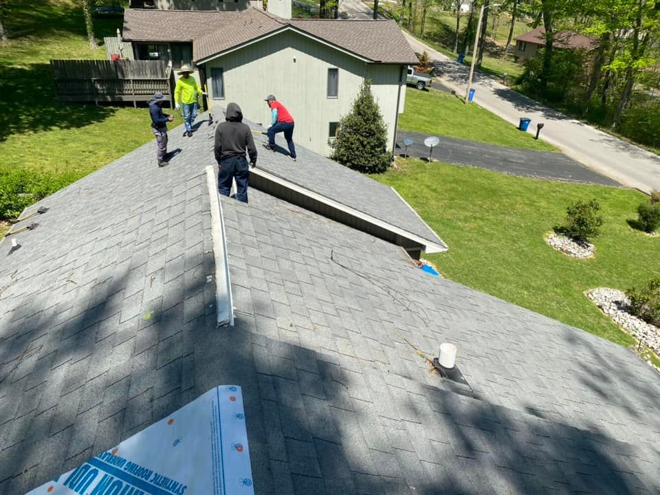 People working on a gray shingled rooftop near a house on a sunny day