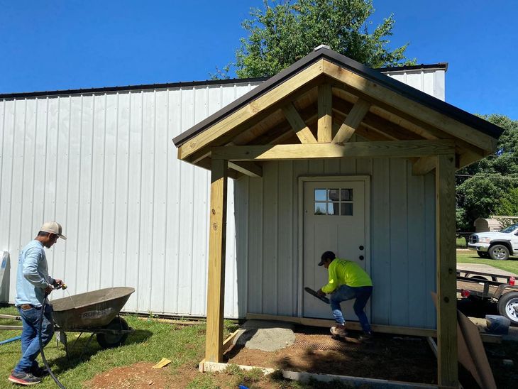 Two workers building a small porch on a beige shed beside a white building.