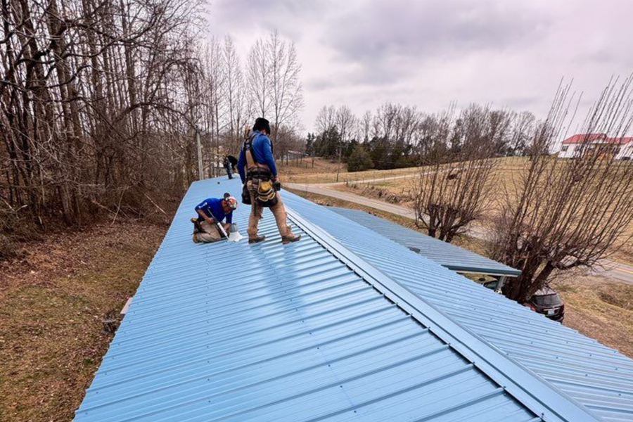 Workers on a blue metal roof, one kneeling and one standing, with trees and fields in the background