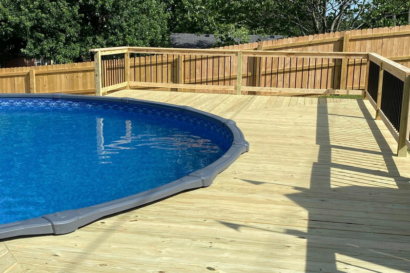 Backyard pool with blue water, wooden deck, and tan fence in bright sunlight