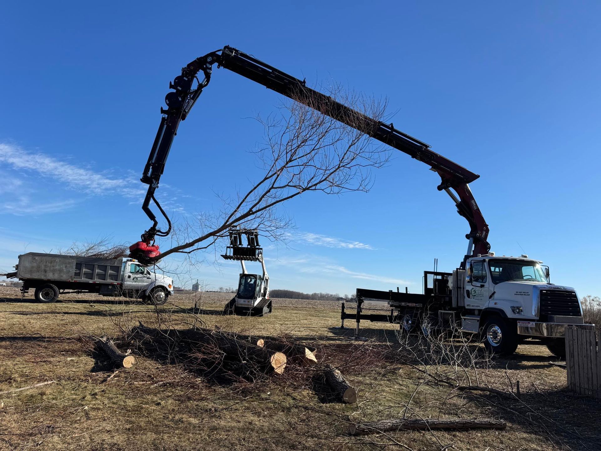 Truck with crane lifting a tree branch into a chip truck under a clear blue sky.