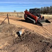 Skid steer grader smoothing a dirt road near a fenced field under a blue sky