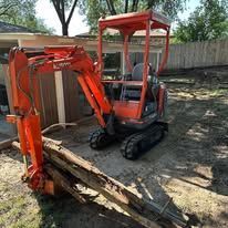 Orange mini excavator with front blade parked in a dirt backyard beside a fence and shed
