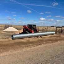 A skid steer carries a long metal pipe on a dirt lot under a blue sky.