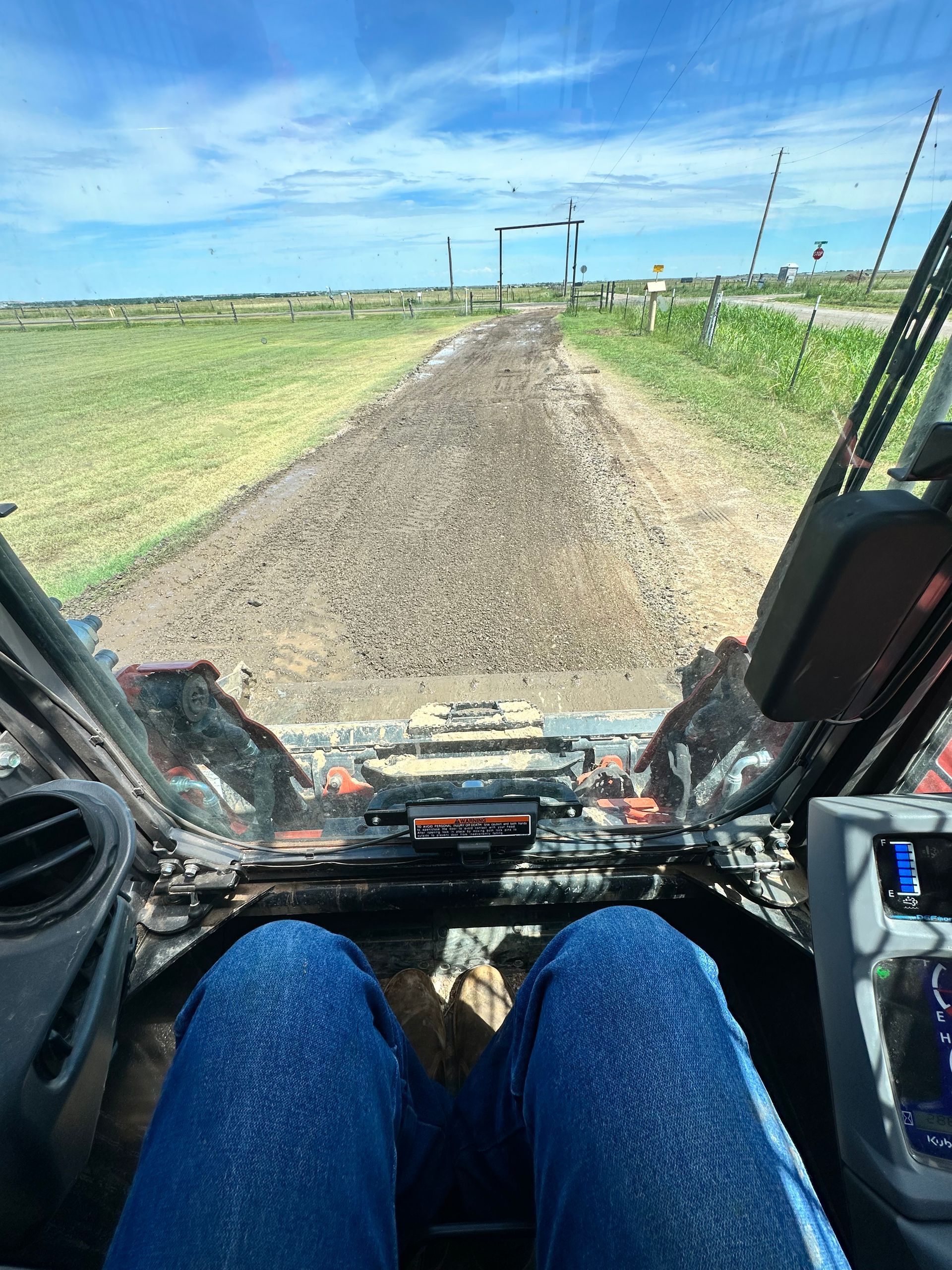 View from a vehicle driving down a gravel farm road through open green fields under a blue sky