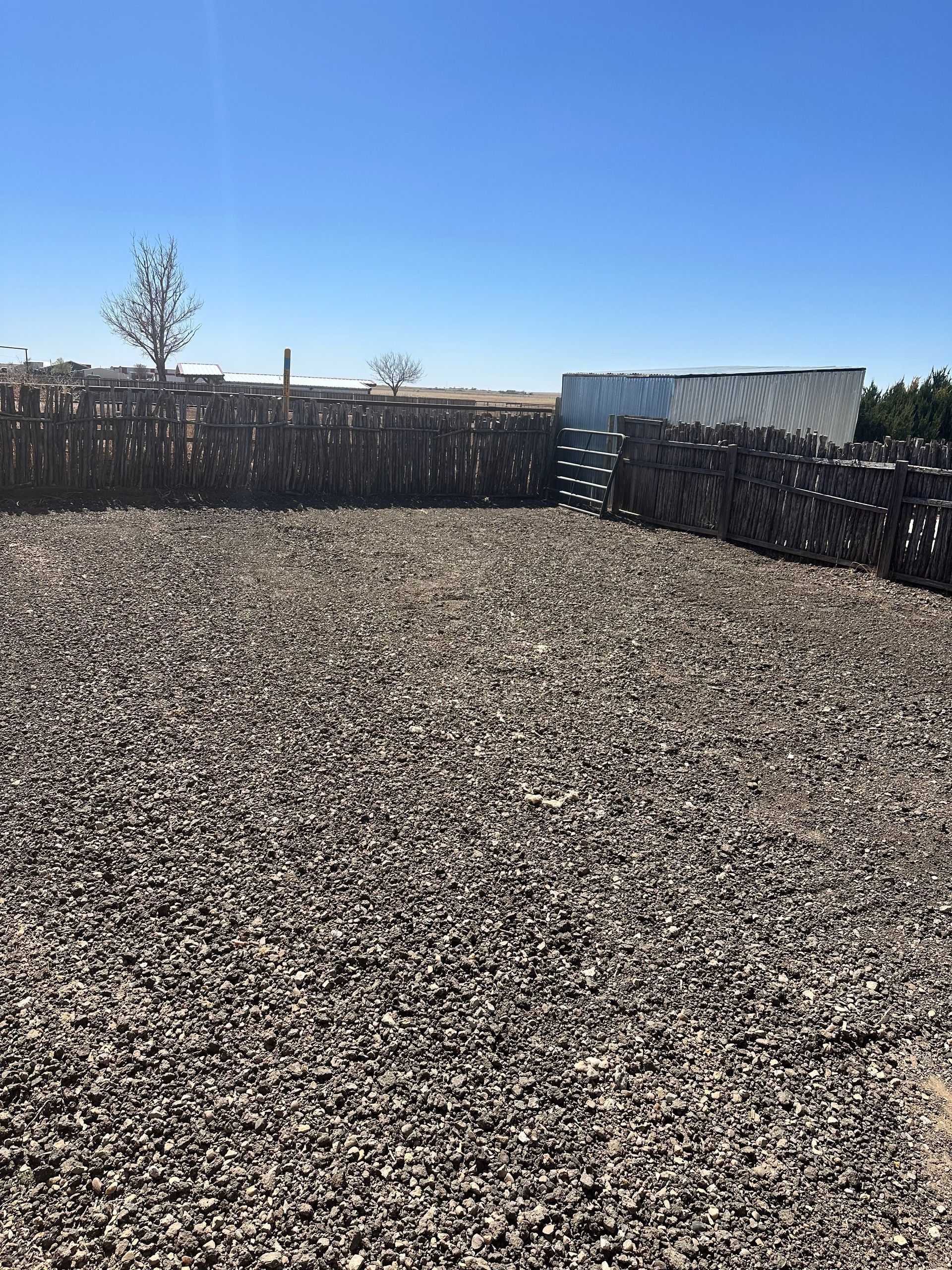 Large gravel lot under a clear blue sky, bordered by dark fences and a few small trees.
