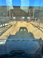 A skid steer loader lifts a long, corrugated metal culvert pipe on a dirt road beside a pile of gravel under a blue sky.