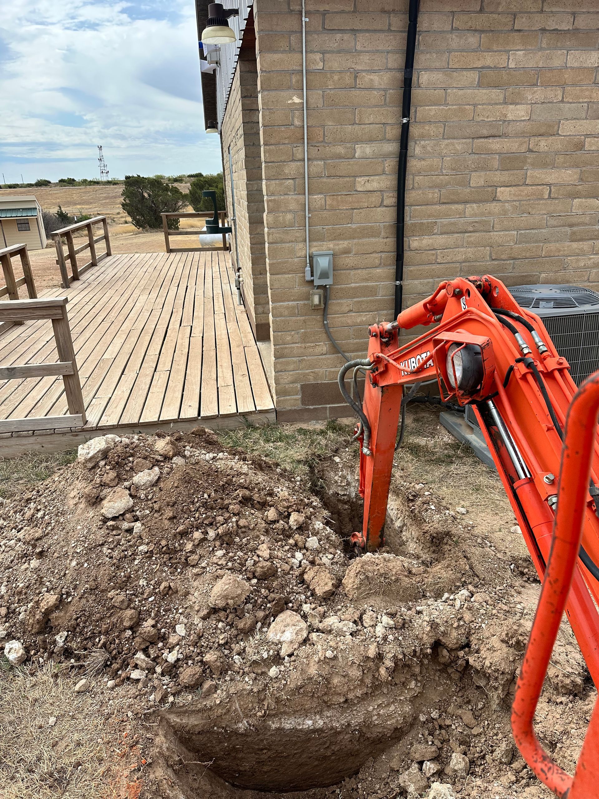 Orange excavator arm digging a hole beside a brick building on a muddy construction site
