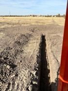 An orange skid-steer loader parked on a dirt road near an exposed black plastic culvert pipe in a rural field.