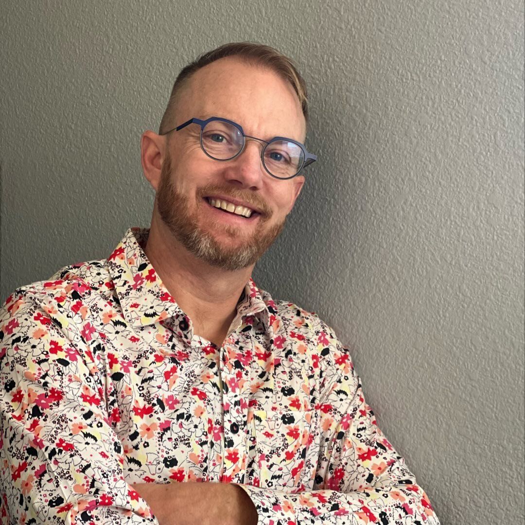 Smiling man with glasses and floral shirt leaning against a gray wall
